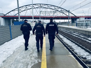 na zdjęciu jeden policjant umundurowany idący z dwoma strażnikami ochrony kolei po peronie
