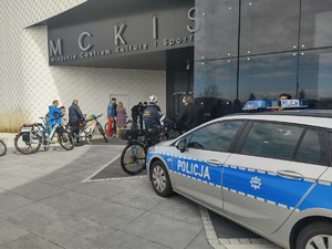 na zdjęciu znajduje się parking przed halą sportową Miejskie Centrum Kultury i Sportu, radiowóz policyjny oraz osoby z rowerami, strażnik miejski