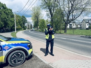 na zdjęciu umundurowany policjant ruchu drogowego stojący obok radiowozu policyjnego dokonuje pomiaru prędkości na drodze